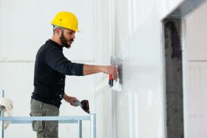 Drywall Worker in Yellow Helmet Plastering Wall While Standing on Scaffold with Workers Comp Insurance in Columbia